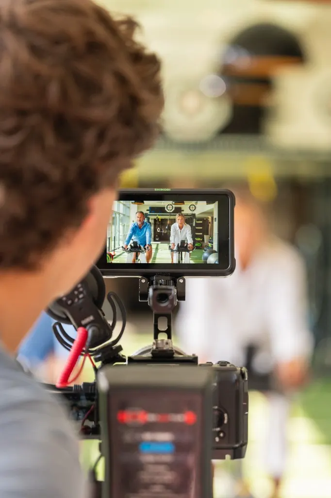 Over-the-shoulder view of a camera monitor showing two people on exercise bikes